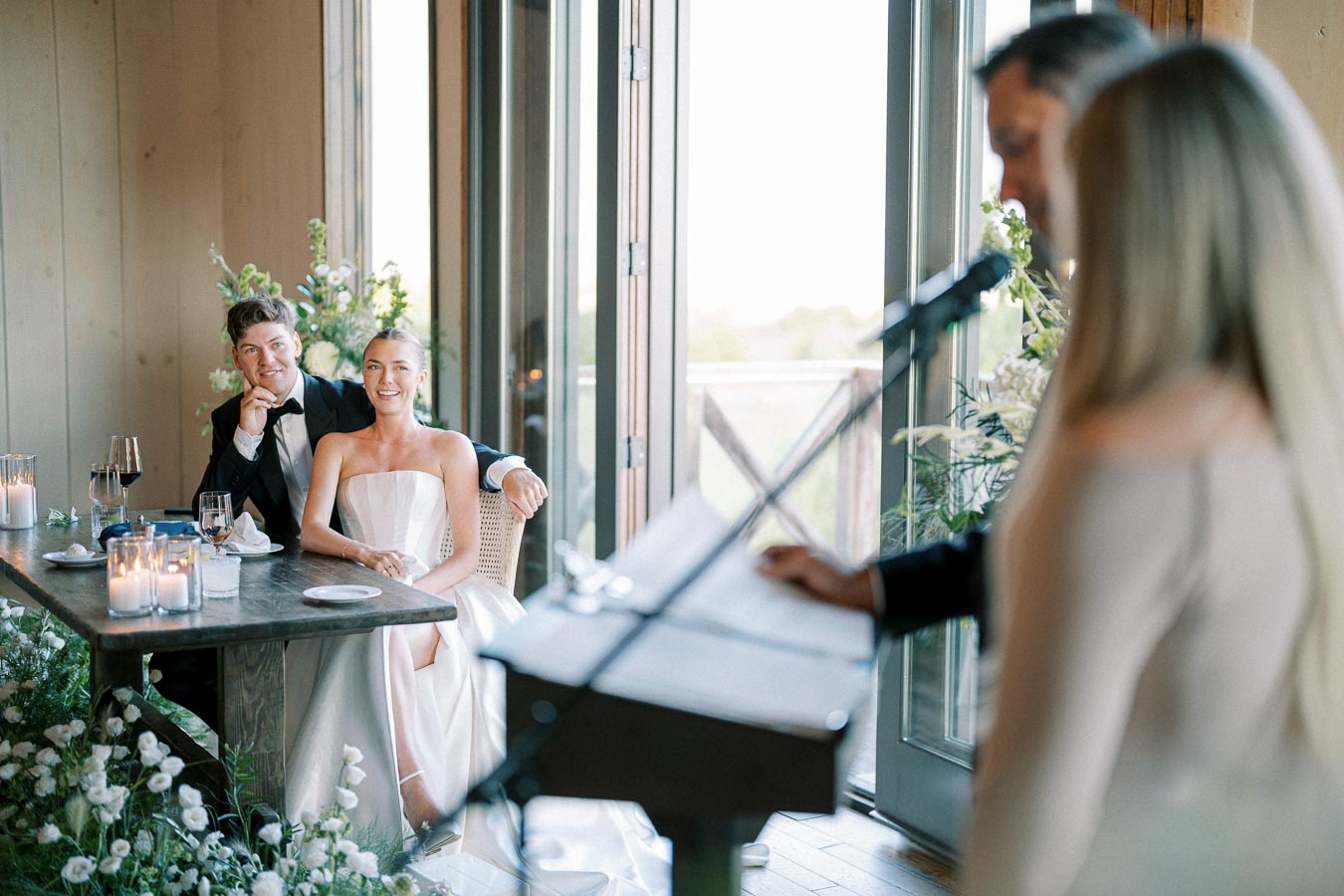 A bride and groom sit together at a decorated table, smiling during a speech at a wedding reception. Candles, flowers, and a large window with a scenic view create a romantic ambiance, while guests speak at the podium.