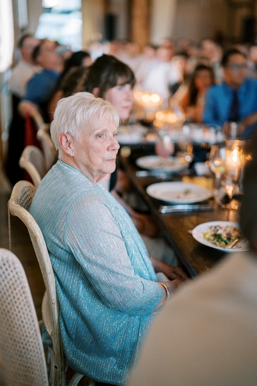 Elderly woman in a blue jacket sitting at a dining table during a social event, surrounded by other attendees with plates of food and candlelit ambiance.