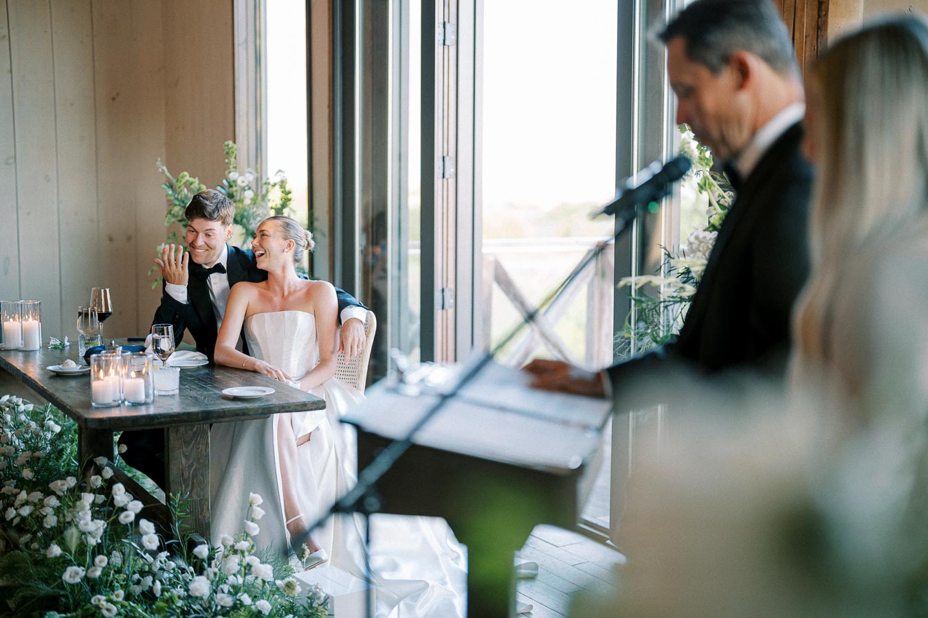 A bride and groom sitting at a decorated wedding reception table, smiling and enjoying a speech from a man with a microphone, surrounded by flowers and candlelight.