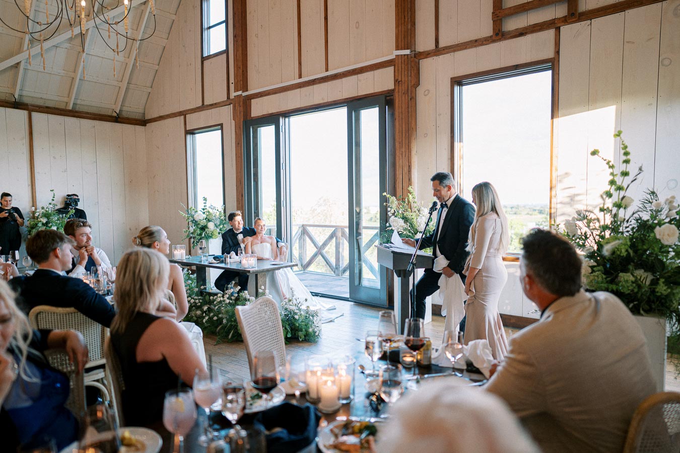 Wedding reception in a rustic venue with guests seated, focusing on a couple at the head table listening to a speech, surrounded by elegant floral arrangements and candlelit tables.