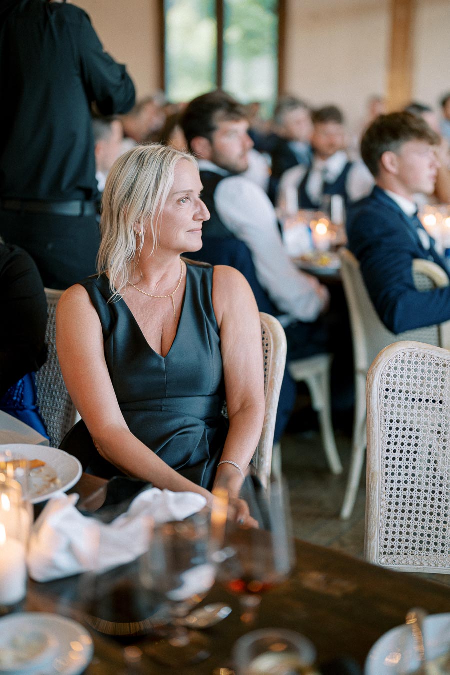 A woman in a formal dark dress sits at a banquet table, surrounded by elegantly dressed guests at a wedding reception, with soft lighting and candles creating a warm ambiance.