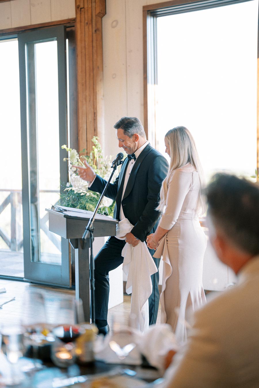 A man in a tuxedo gives a speech at a wedding reception, standing at a podium with a microphone. A woman in a formal beige dress stands beside him, while guests watch from a table in the foreground. The room features large windows and elegant decor.