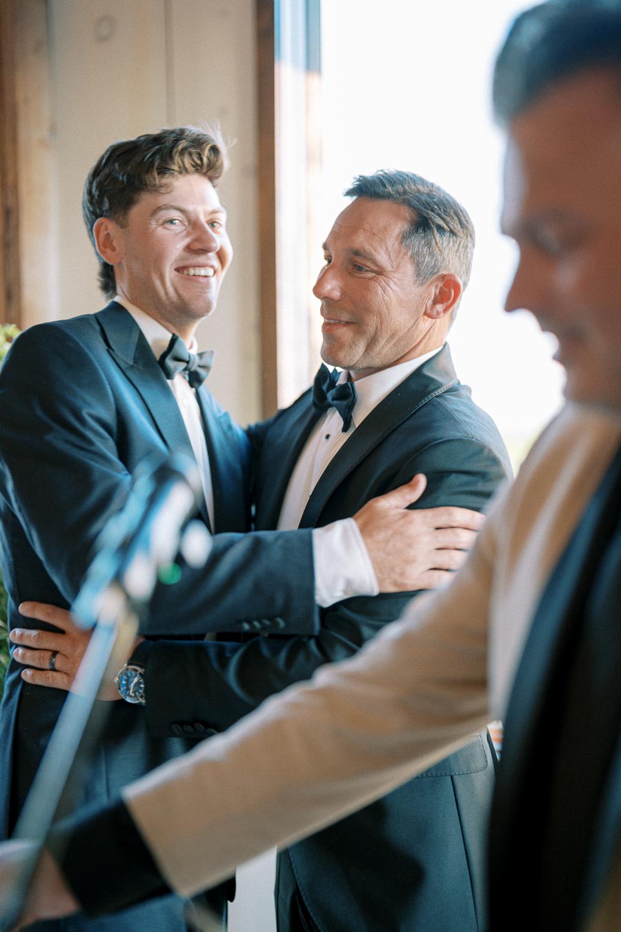 Two men in elegant tuxedos embrace warmly at a formal event, with one smiling broadly, capturing a joyful and celebratory moment. A person in partial view is seen in the foreground.