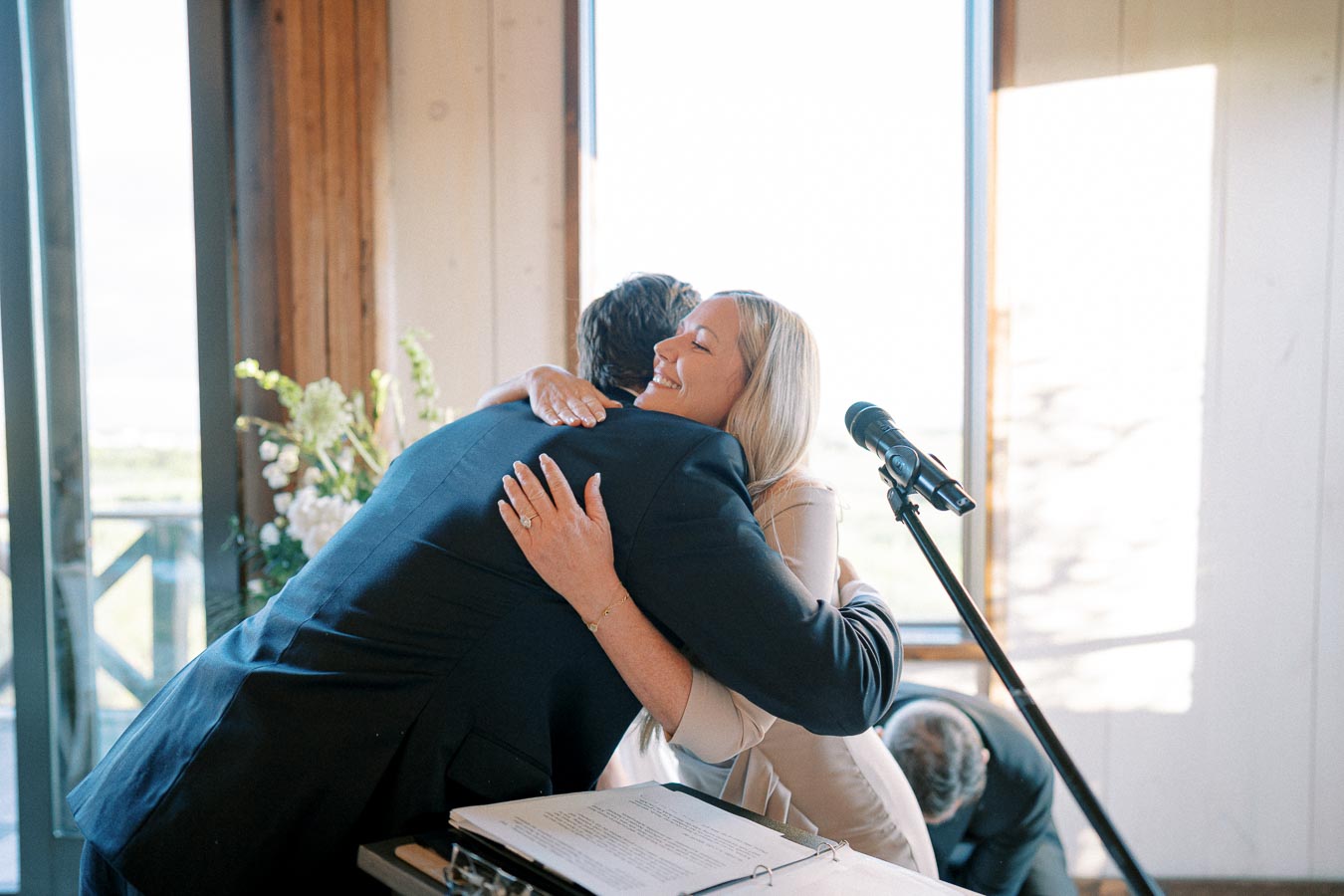 A woman and man exchanging a warm hug at a podium during a formal event, with a microphone in the foreground and natural light streaming through large windows.