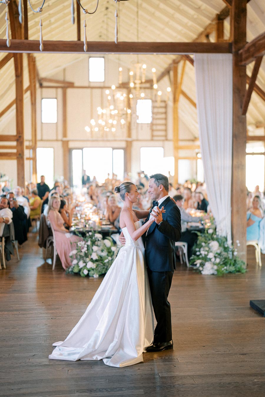 Father and bride sharing a dance in a beautifully decorated rustic wedding venue with guests seated at elegantly adorned tables and soft lighting highlighting the wooden interior.