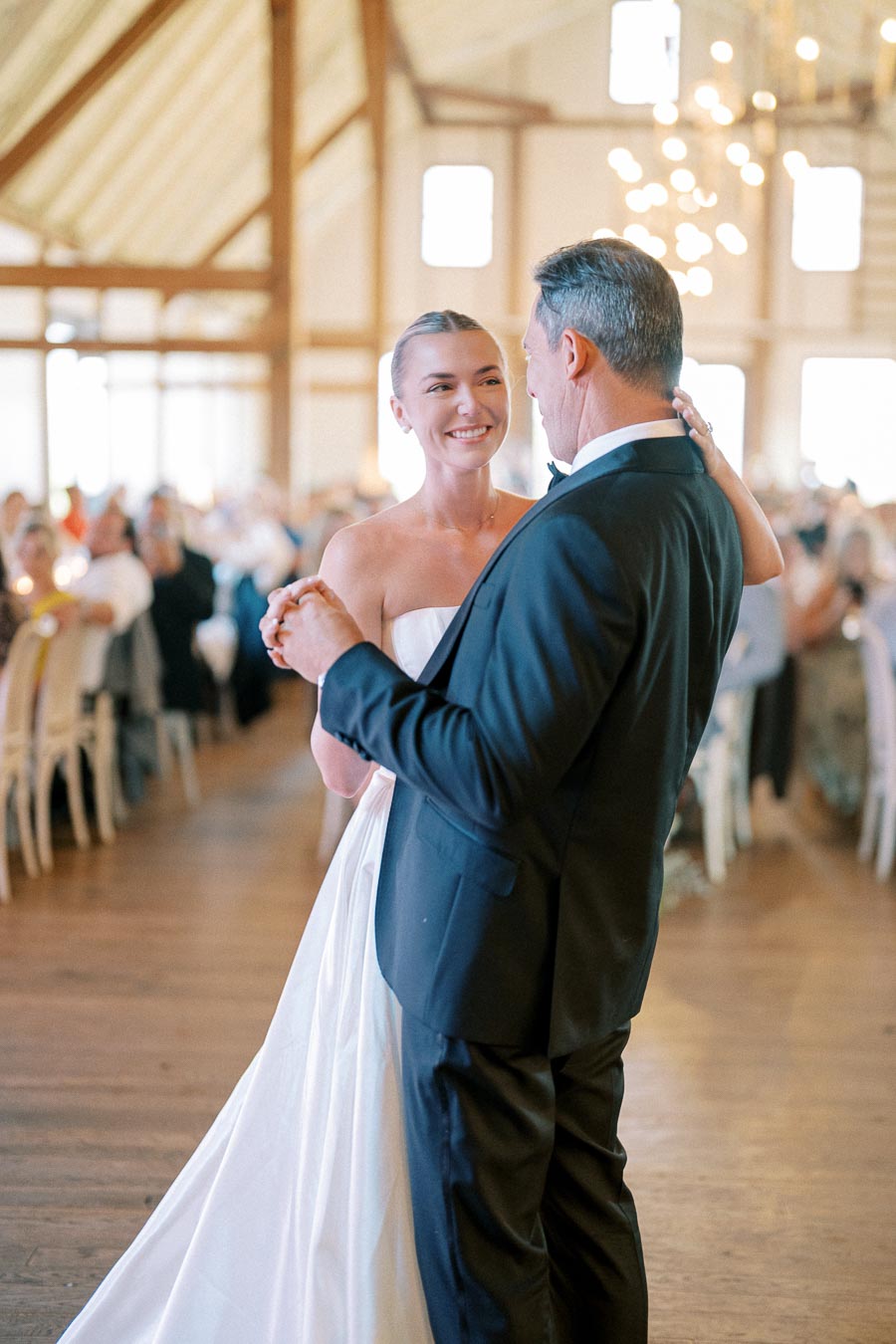 Father and daughter dance at indoor wedding reception, bride in white gown smiling while dancing with well-dressed older man, warm lighting and blurred guests in elegant venue.