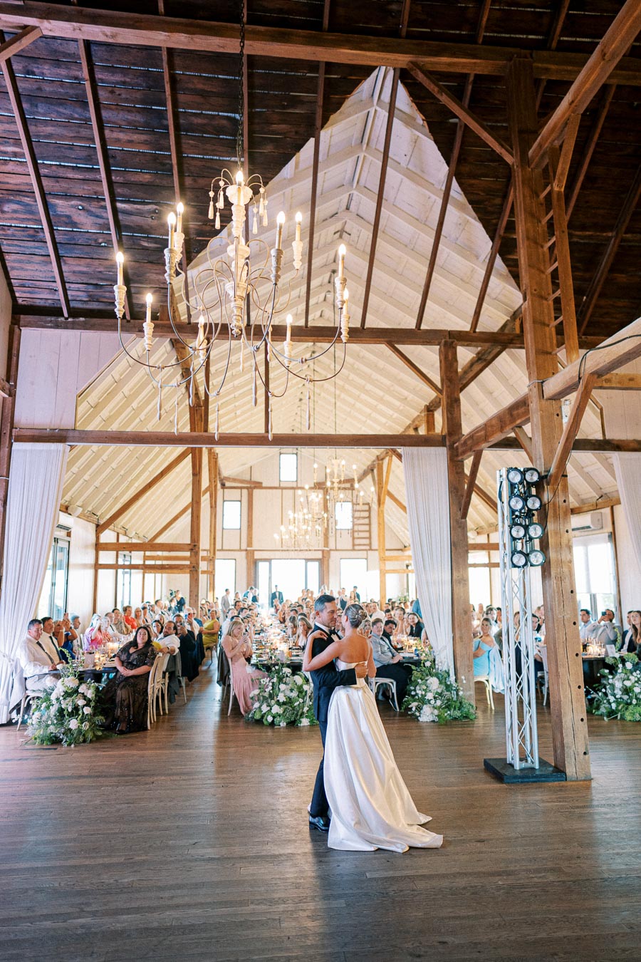 A couple shares a romantic first dance in a rustic, elegantly decorated wedding venue with wooden beams and chandeliers, surrounded by seated guests.