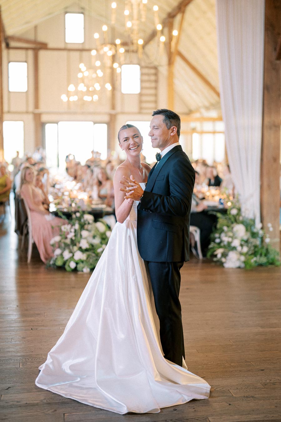 A bride and groom share a joyful first dance in an elegantly decorated barn venue, adorned with chandeliers and floral arrangements, surrounded by guests.