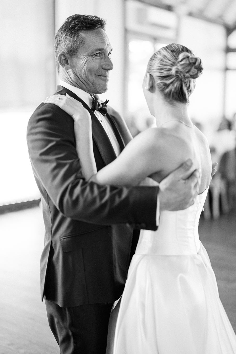 A father and daughter share a joyful dance at a wedding, with the father wearing a tuxedo and the bride in a white gown, in a black and white photograph.