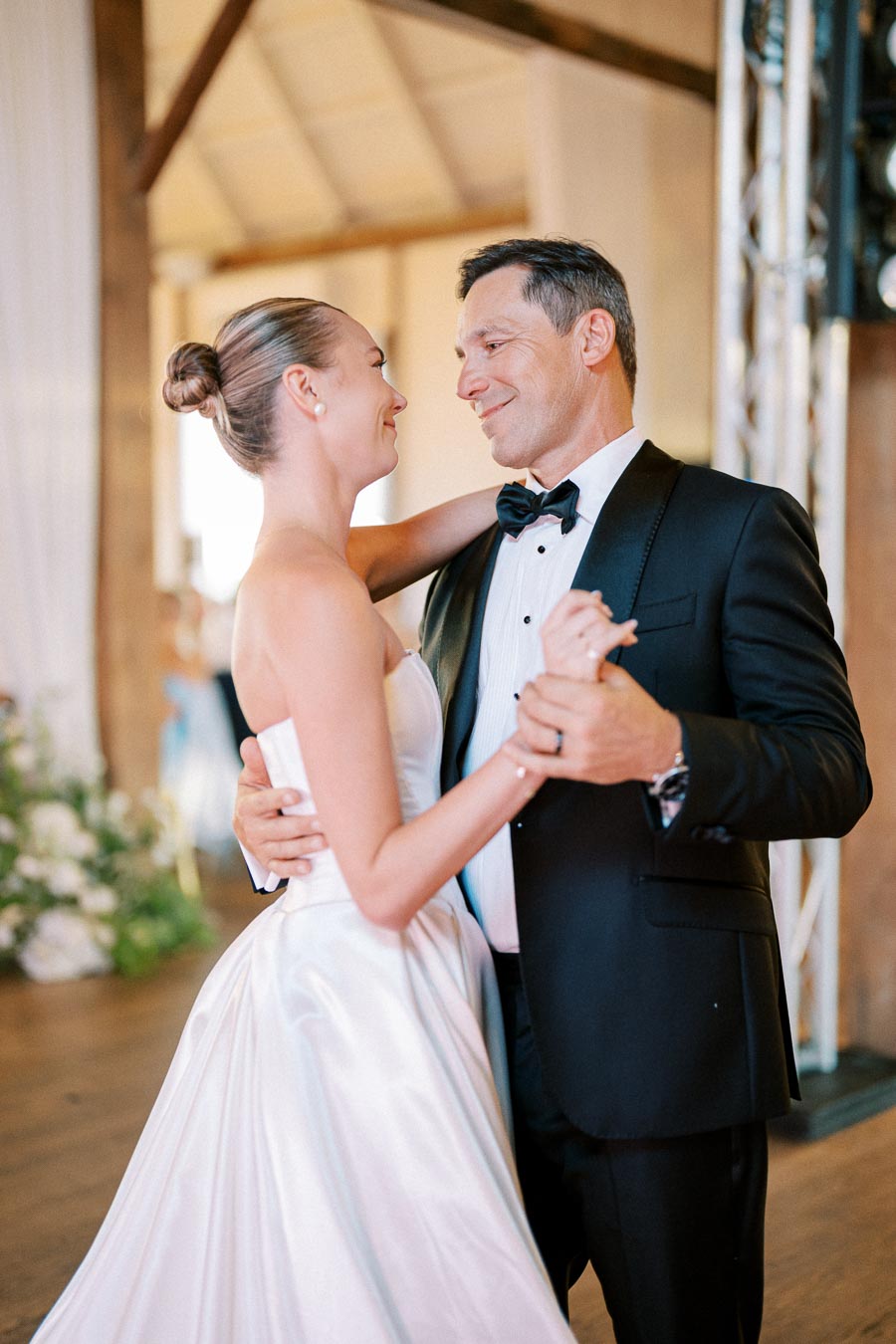 A bride in a white wedding dress and a man in a tuxedo share a dance at a wedding reception, smiling at each other in a warmly lit venue.