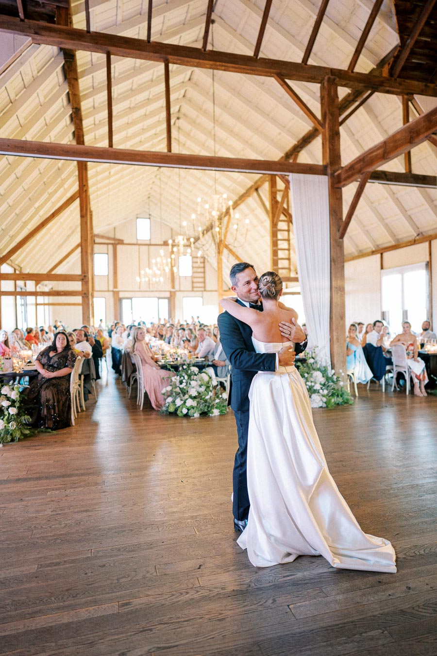 Father and bride share a heartwarming dance in a beautifully decorated rustic barn wedding venue, surrounded by guests seated at elegantly adorned tables.