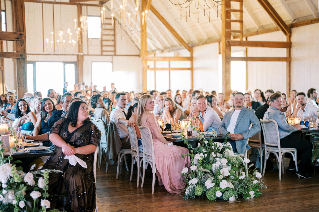 A large group of elegantly dressed guests seated at tables in a rustic, chandelier-lit venue, attentively watching an event or performance.