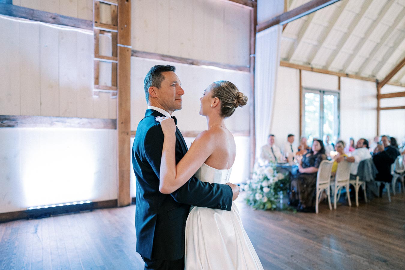 A bride and groom share a first dance in a rustic barn wedding venue, with guests seated at tables in the background. The bride wears an elegant white gown, and the groom is in a classic black tuxedo.