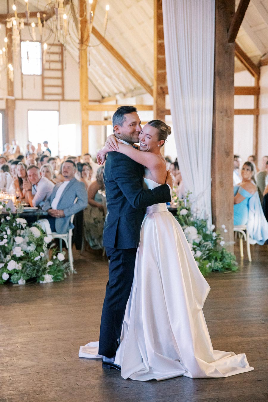 Bride and groom sharing their first dance at a rustic indoor wedding reception, surrounded by guests and floral decorations.