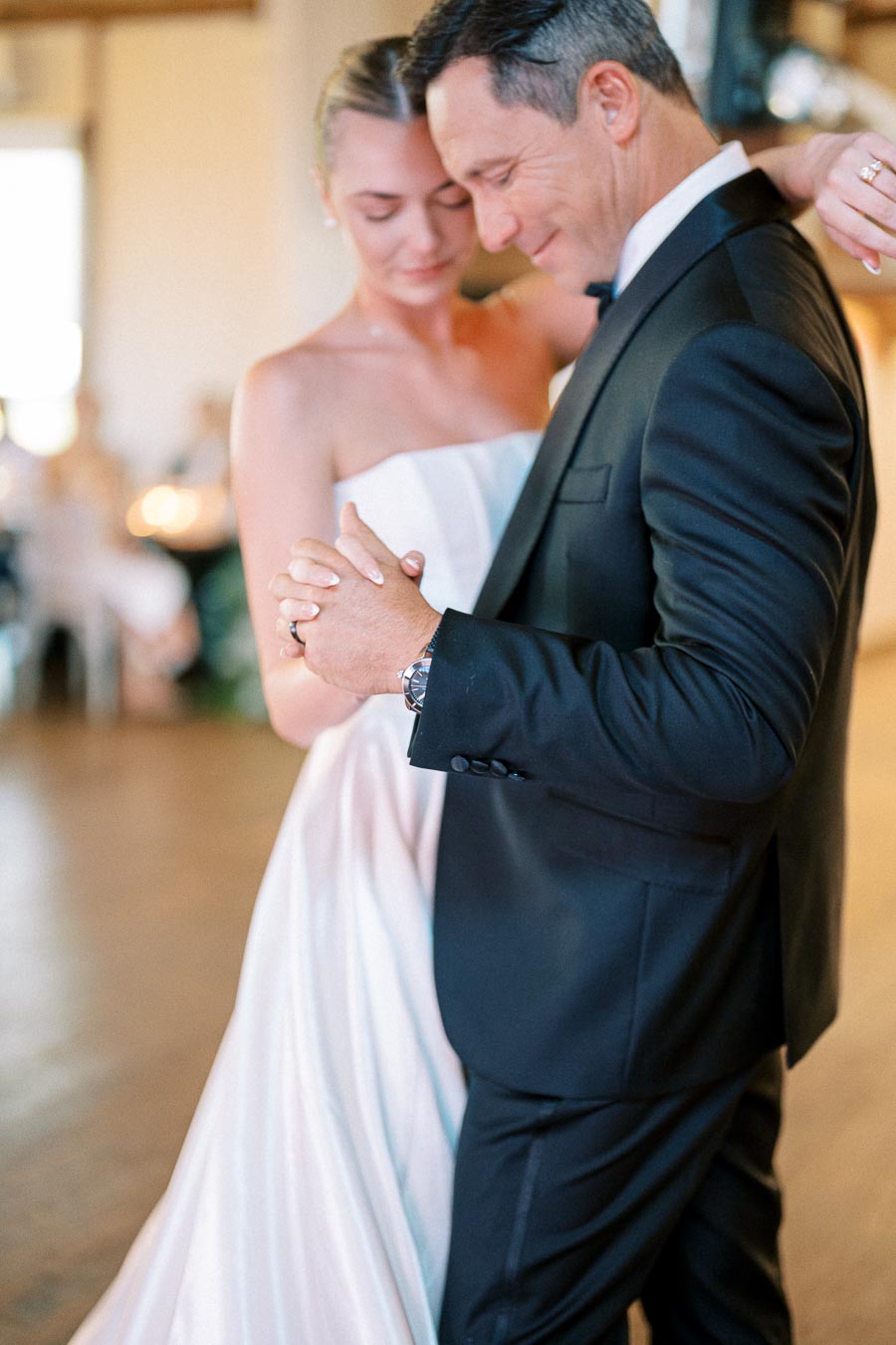 A bride in a white wedding dress shares a dance with a man in a black suit at a wedding reception, highlighting a tender moment on the dance floor.