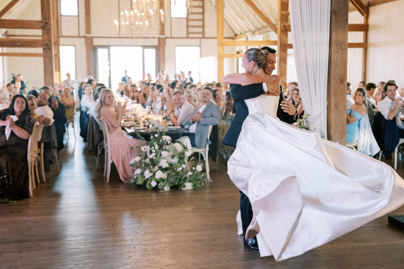 A bride and groom share a joyful dance at their wedding reception, surrounded by applauding guests in a beautifully decorated rustic venue with elegant chandeliers and floral arrangements.