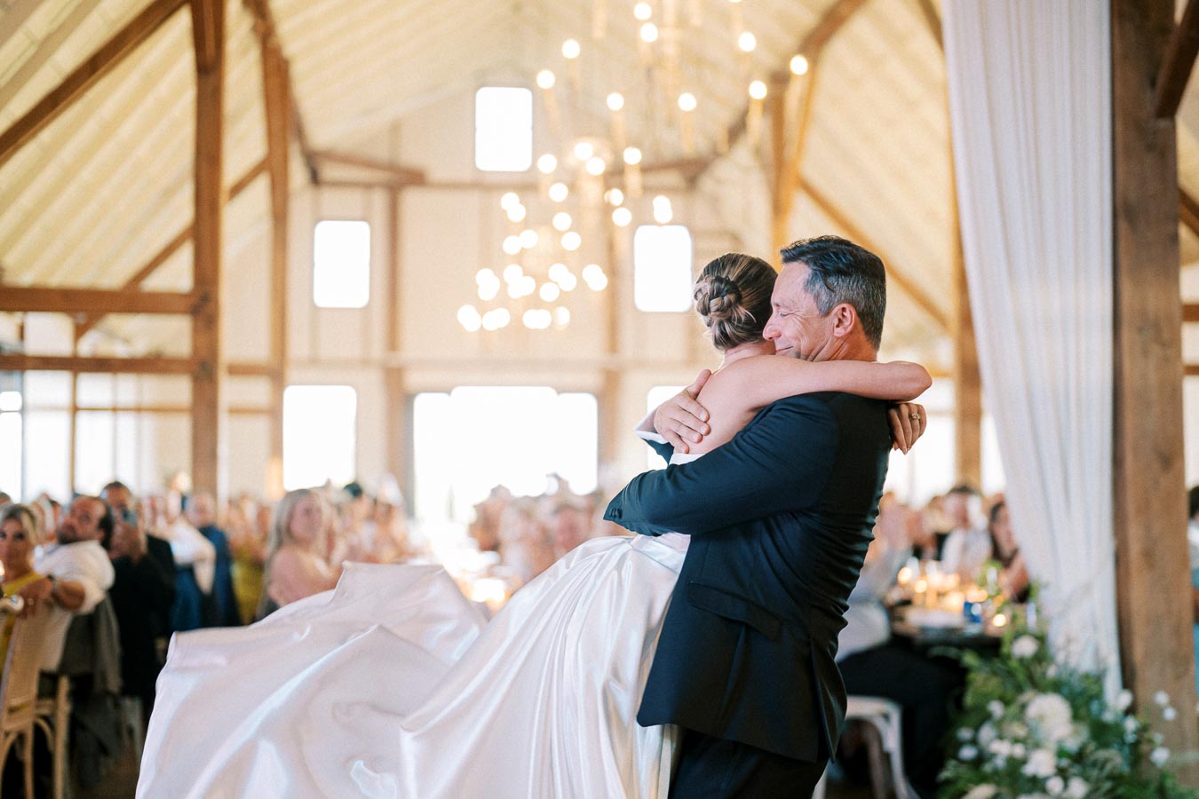Father and bride embrace during emotional dance at a wedding reception in a beautifully decorated barn venue, surrounded by guests.