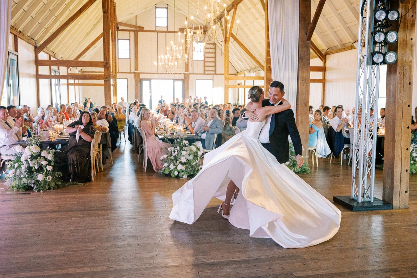 A bride and groom share their first dance in a beautifully decorated rustic barn wedding venue, surrounded by guests clapping and seated at elegantly set tables.