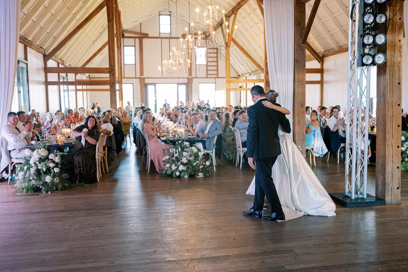 A couple sharing a dance at a rustic wedding reception in a beautifully decorated barn, surrounded by seated guests applauding and enjoying the celebration.