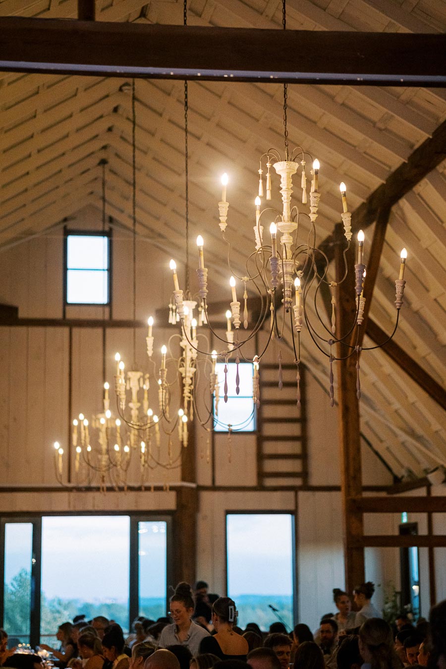 Elegant chandeliers with glowing candles illuminate a rustic barn wedding reception, filled with guests enjoying the celebration.