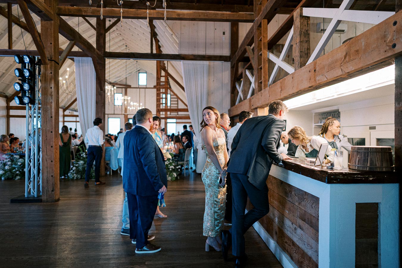 People socializing and ordering drinks at a rustic barn wedding reception, featuring wood beams and elegant decor.