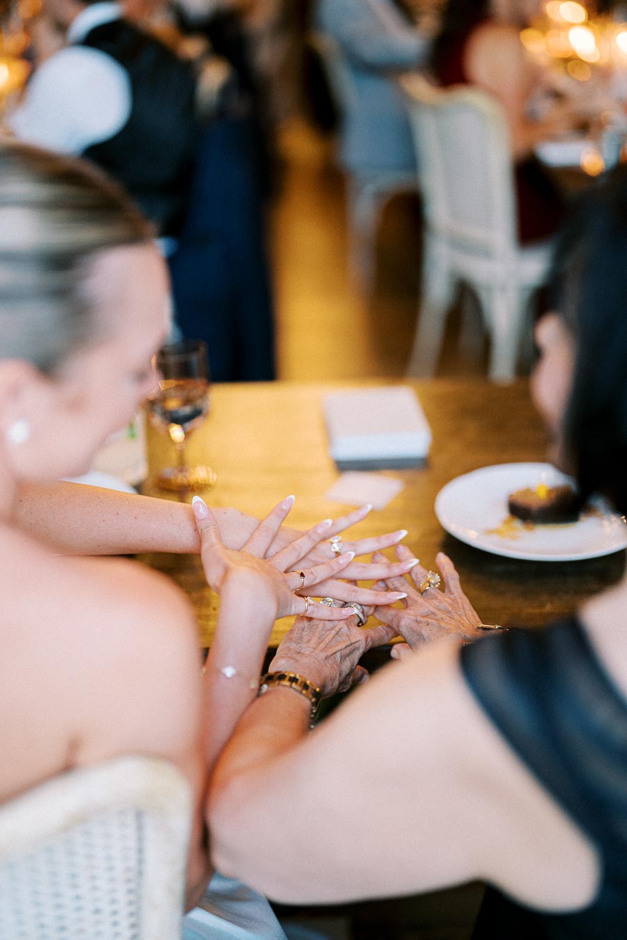 A close-up of two women at a wedding reception, showing their hands adorned with elegant jewelry and rings, sharing a moment of joy and connection at a table set with a dessert plate.