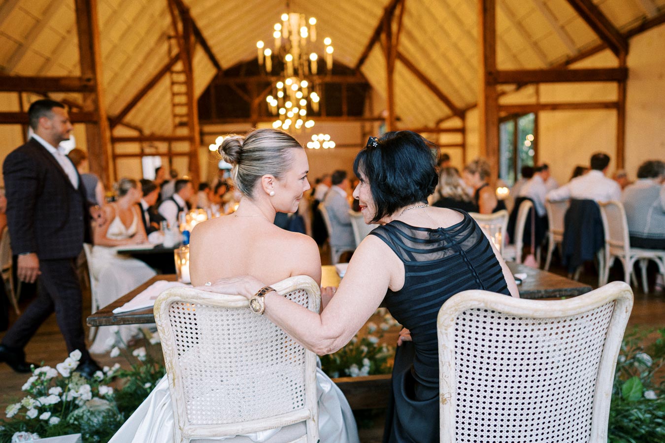 Wedding reception in rustic venue, featuring two women seated on elegant chairs, embracing and smiling amid warmly lit chandeliers and well-dressed guests.