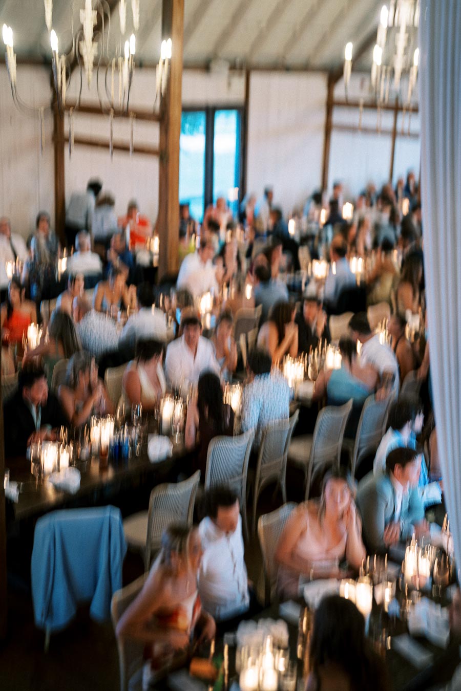 A crowded dining hall with tables filled with guests dressed in formal attire, ambient candlelight creating a warm and inviting atmosphere, and elegant chandeliers overhead.