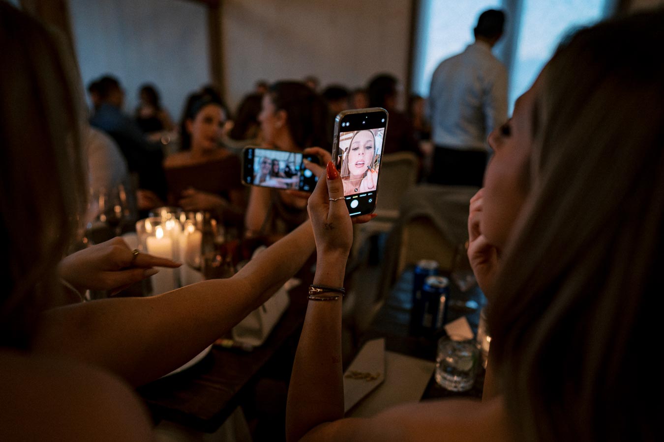 A group of people at a dimly lit dinner party, with one person capturing a selfie on a smartphone, candles flickering on the table.