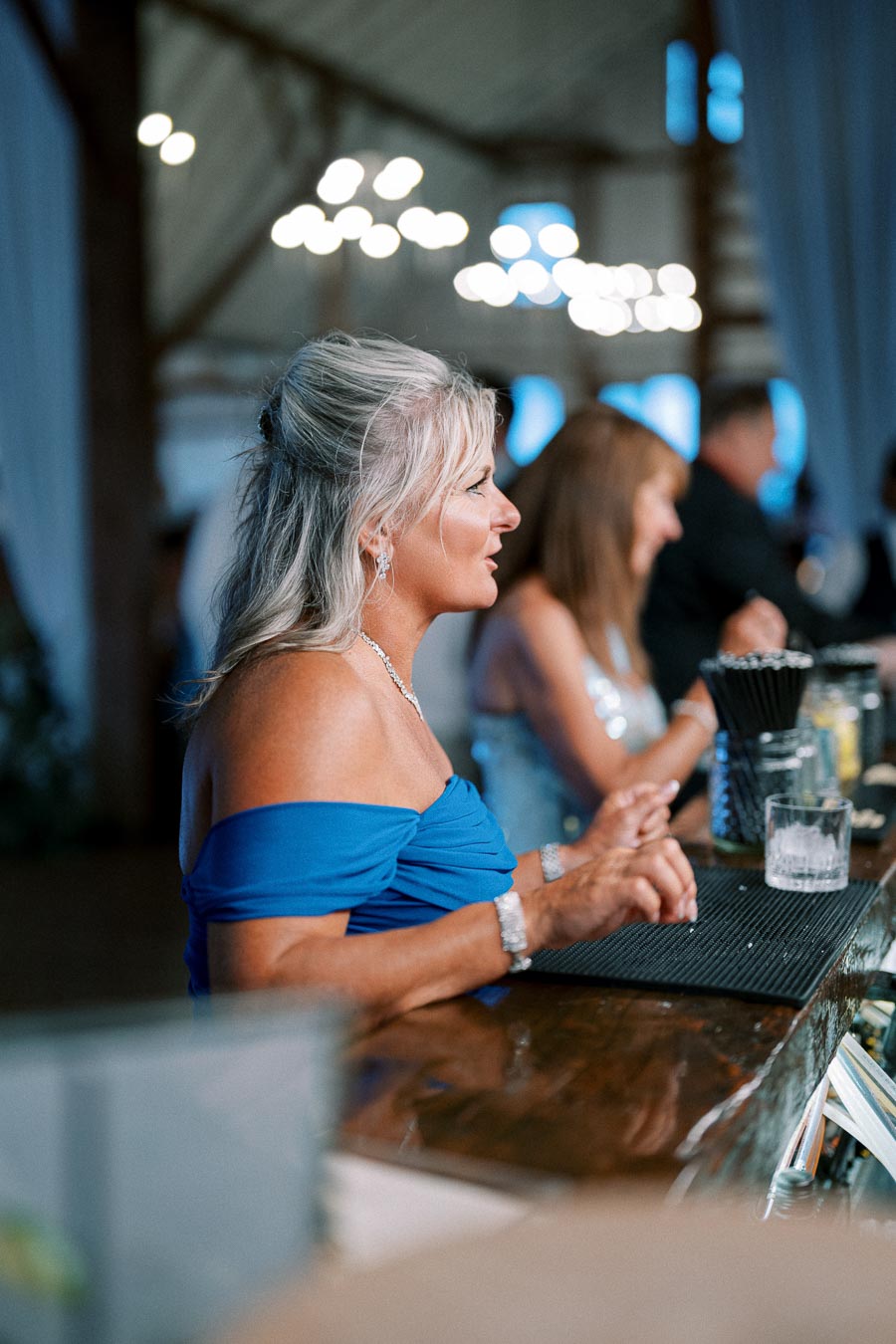 A woman in a blue off-the-shoulder dress sitting at a bar, engaging in conversation, with a blurred background of people and lights in a warmly lit venue.