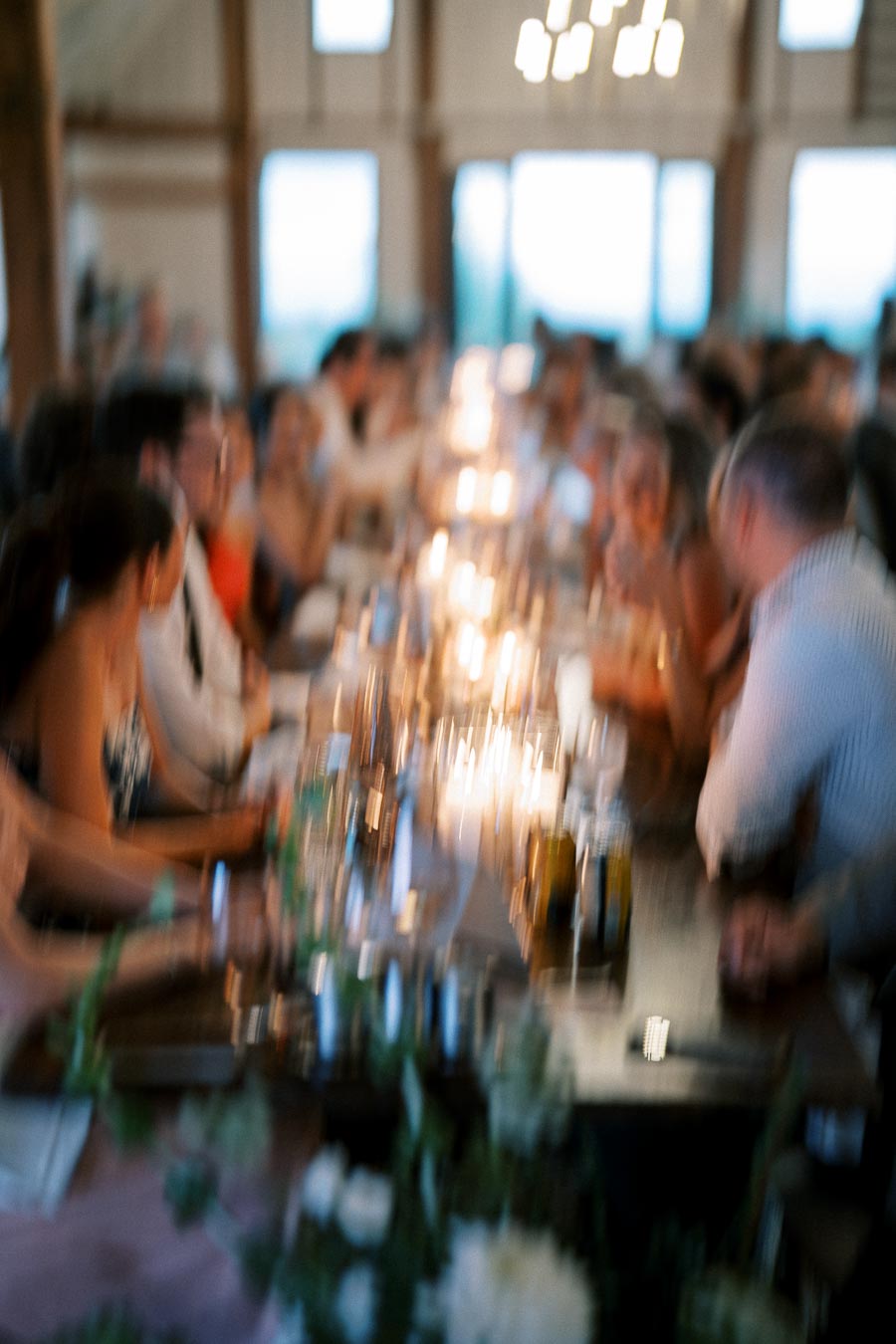 Blurred image of a group of people sitting at a long dining table in a warmly lit indoor setting, enjoying a social gathering or dinner party.