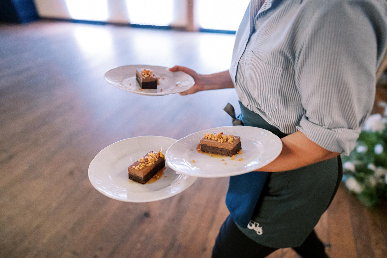 Server carrying three plates of gourmet chocolate dessert garnished with nuts, in a well-lit dining area.