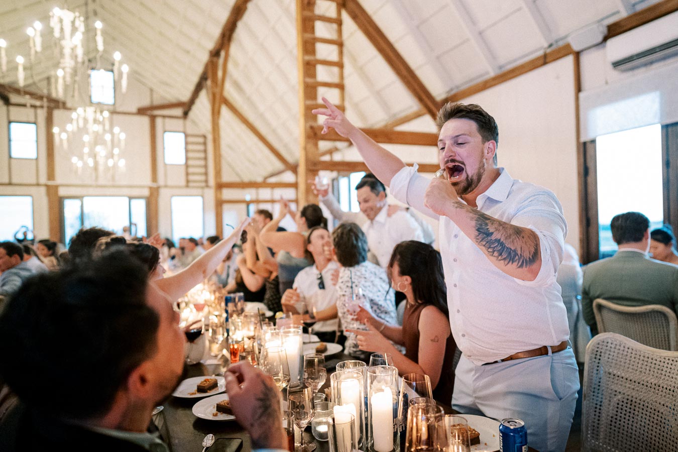 A lively wedding reception in a beautifully decorated barn, featuring a man cheering enthusiastically at a table filled with guests and lit candles, creating a joyful and festive atmosphere.