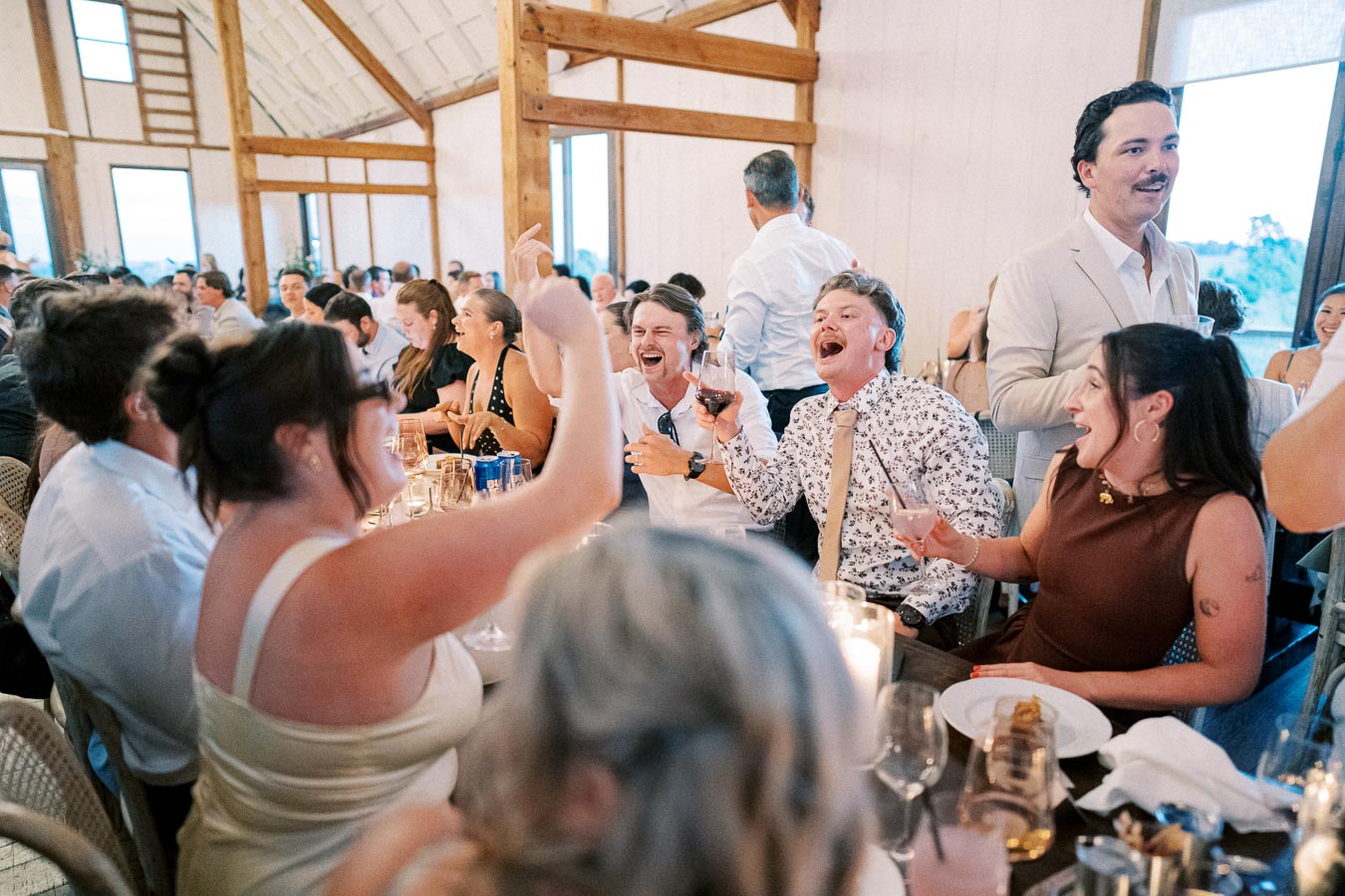 Group of people joyfully celebrating at a lively indoor wedding reception, raising glasses and laughing around a beautifully decorated table with candles and elegant tableware.