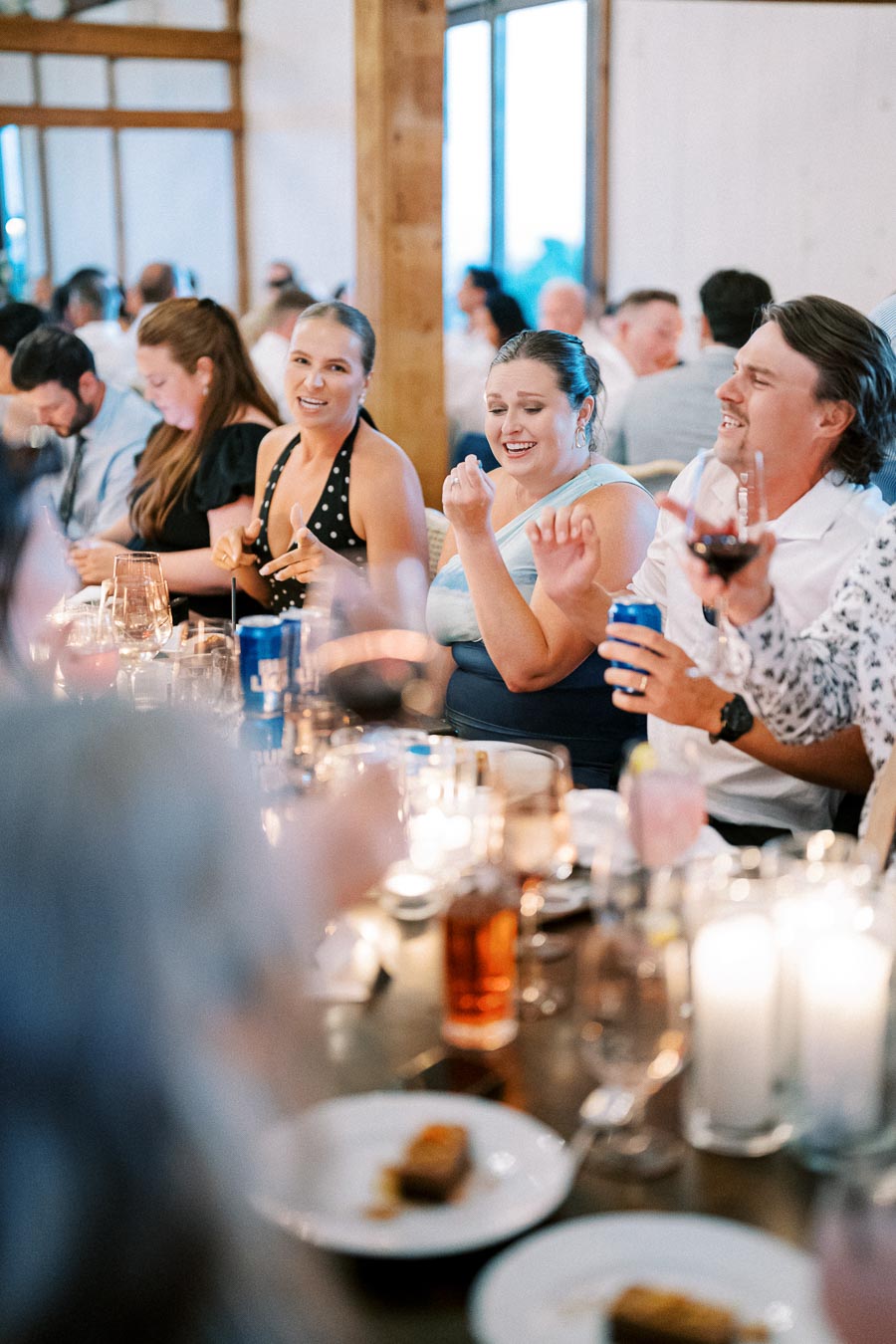 A lively group of people enjoying a wedding reception, seated around a beautifully decorated dinner table with candles and drinks, engaged in conversation and laughter.