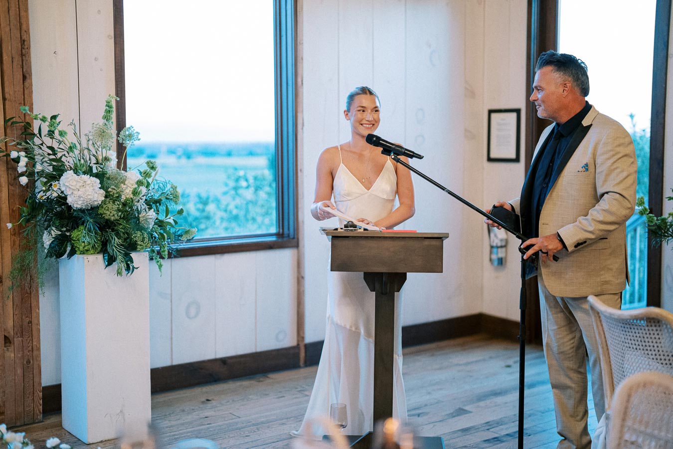 A woman in a white dress stands at a lectern with a microphone, smiling, as a man in a beige suit holds another microphone and listens. They are inside a room with a large window and floral decorations.