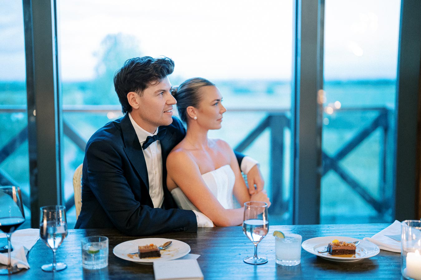 A couple sitting together at a table, elegantly dressed in formal attire, at a scenic venue with large windows overlooking a serene outdoor view.