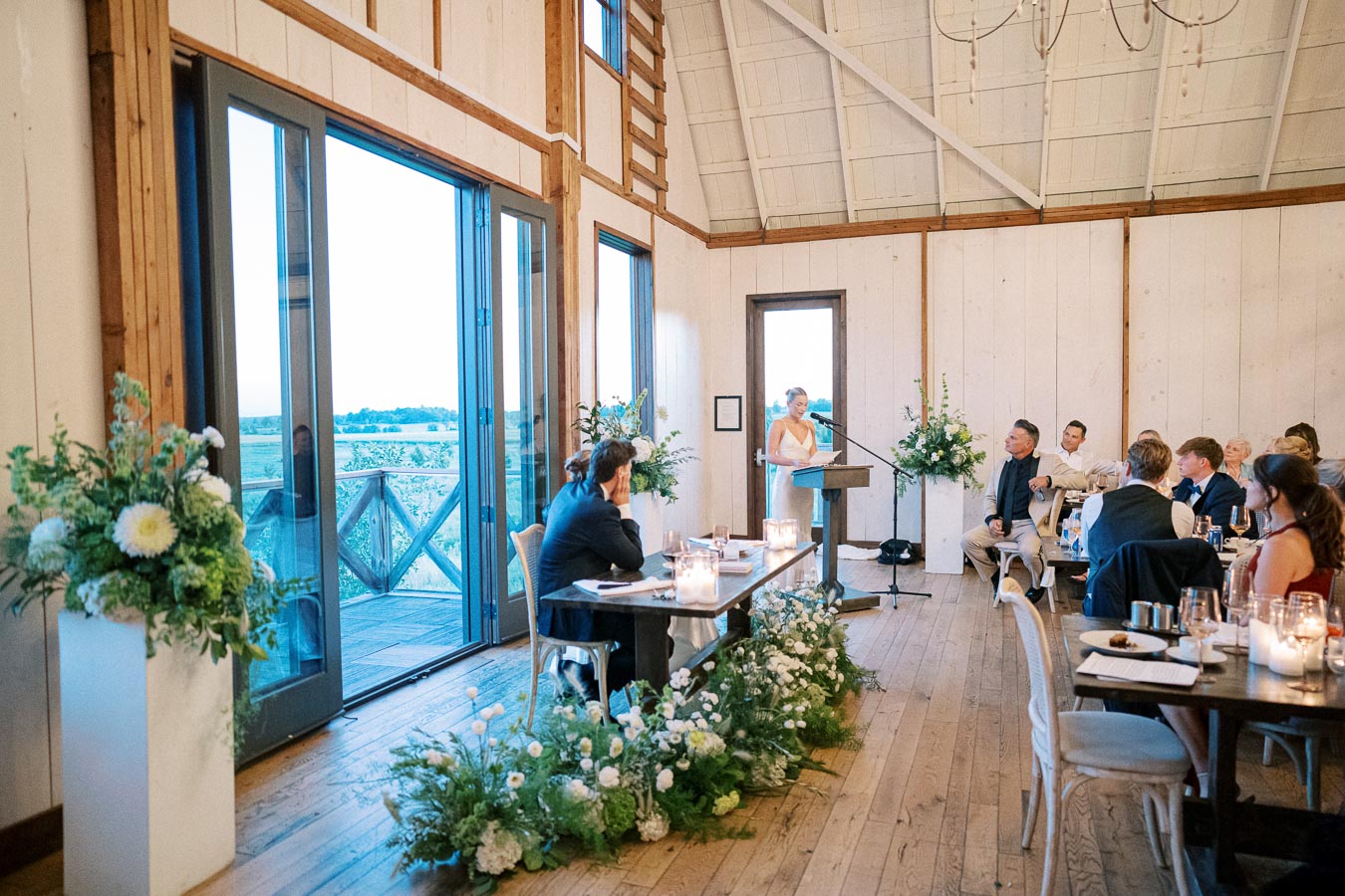 Elegant wedding reception in a rustic venue with wooden floors and large glass doors, featuring a speaker at a podium, surrounded by lush floral arrangements and attentive guests seated at decorated tables.