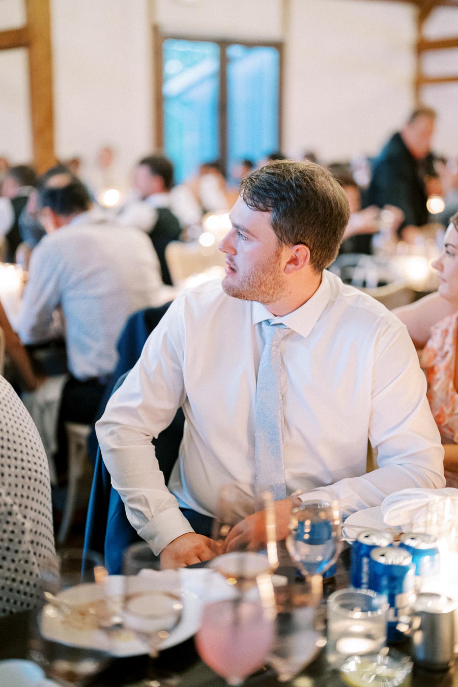 A man in a white dress shirt and tie sitting at a table with drinks and plates during an indoor social event.