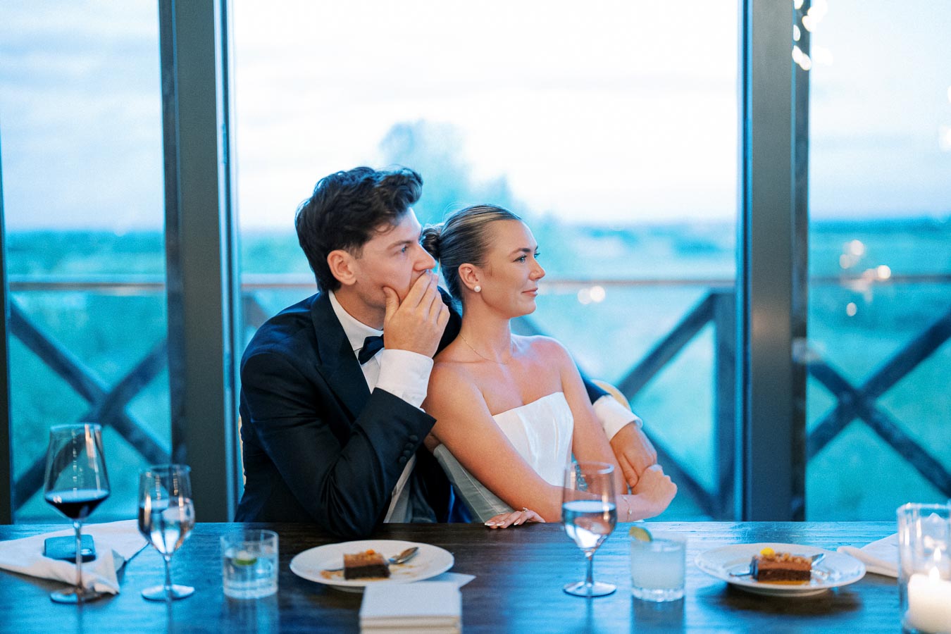 A couple in formal attire enjoying a dinner with scenic views at a restaurant, featuring elegant table settings with wine glasses and plates of dessert.