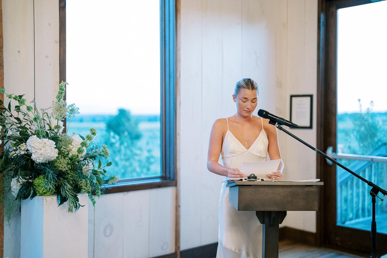 A woman in a white dress stands at a podium inside a venue, reading from a sheet of paper while speaking into a microphone. There is a large vase of white and green flowers beside her, and a large window with a view of the outdoors in the background.