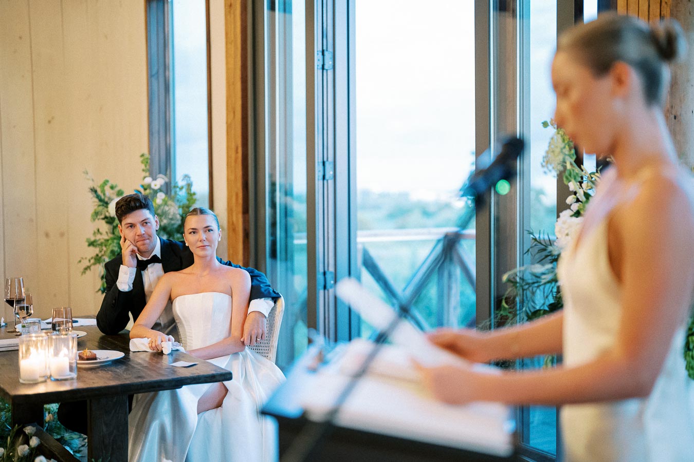 Bride and groom sitting at a table during wedding reception, listening attentively to a speech being delivered by a guest at a microphone, with soft natural lighting and elegant decor in the background.