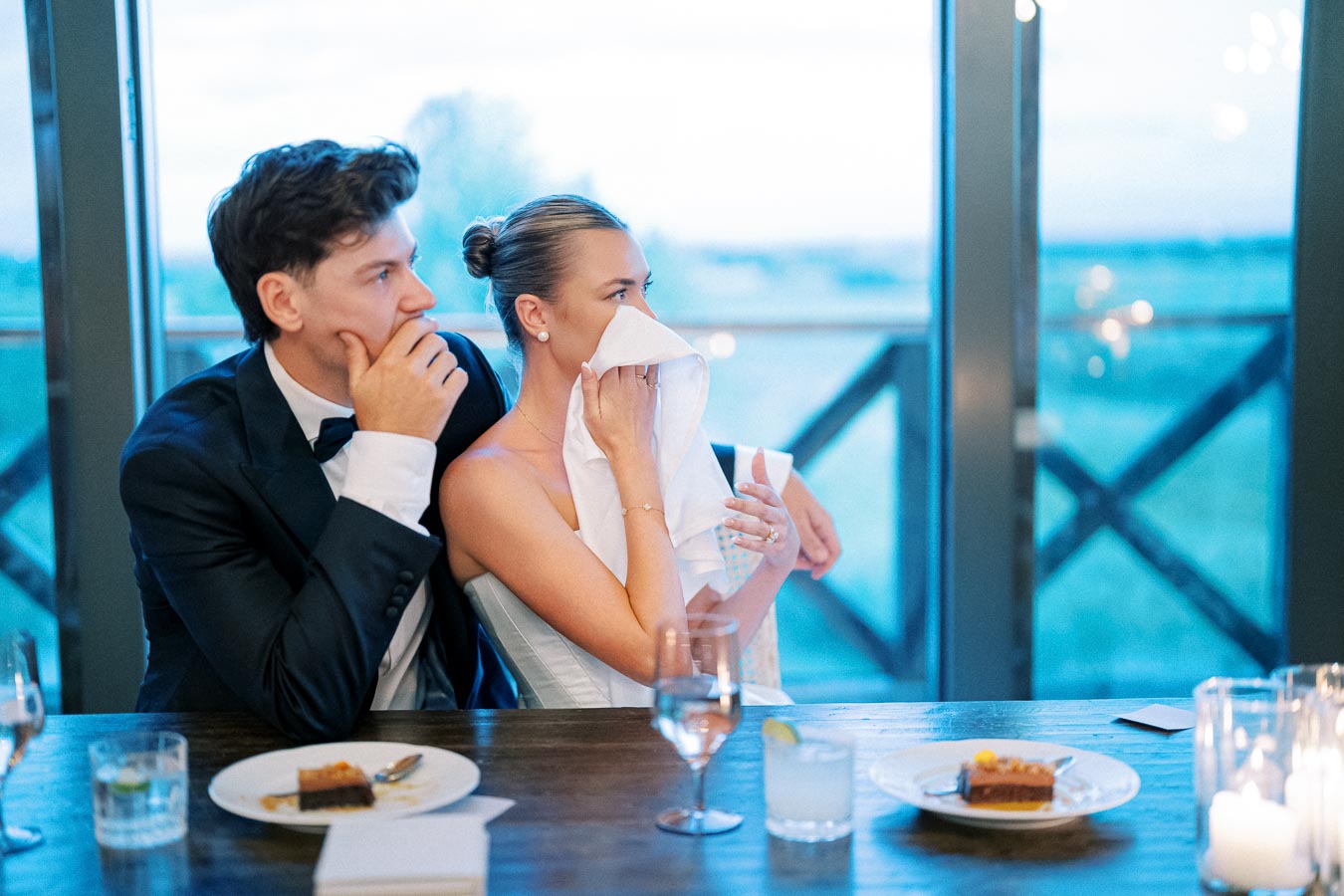A bride wipes away tears with a napkin during a wedding reception, sitting beside a man in a black tuxedo at a wooden table set with half-eaten desserts, drinks, and lit candles.