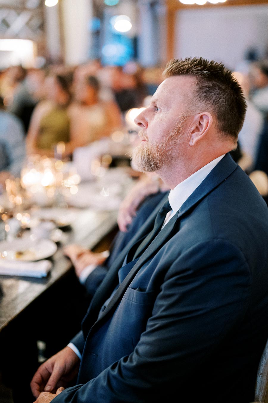 A man in a suit attending a formal event, seated at a table with blurred background, capturing the ambiance of a social gathering.