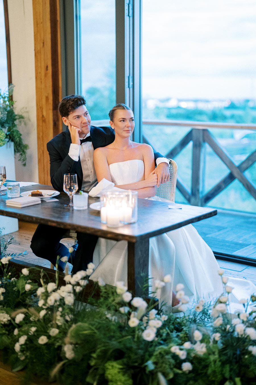 A bride and groom sitting at a rustic wooden table during their wedding reception, surrounded by elegant floral decorations and lit candles, with a scenic view through large windows in the background.