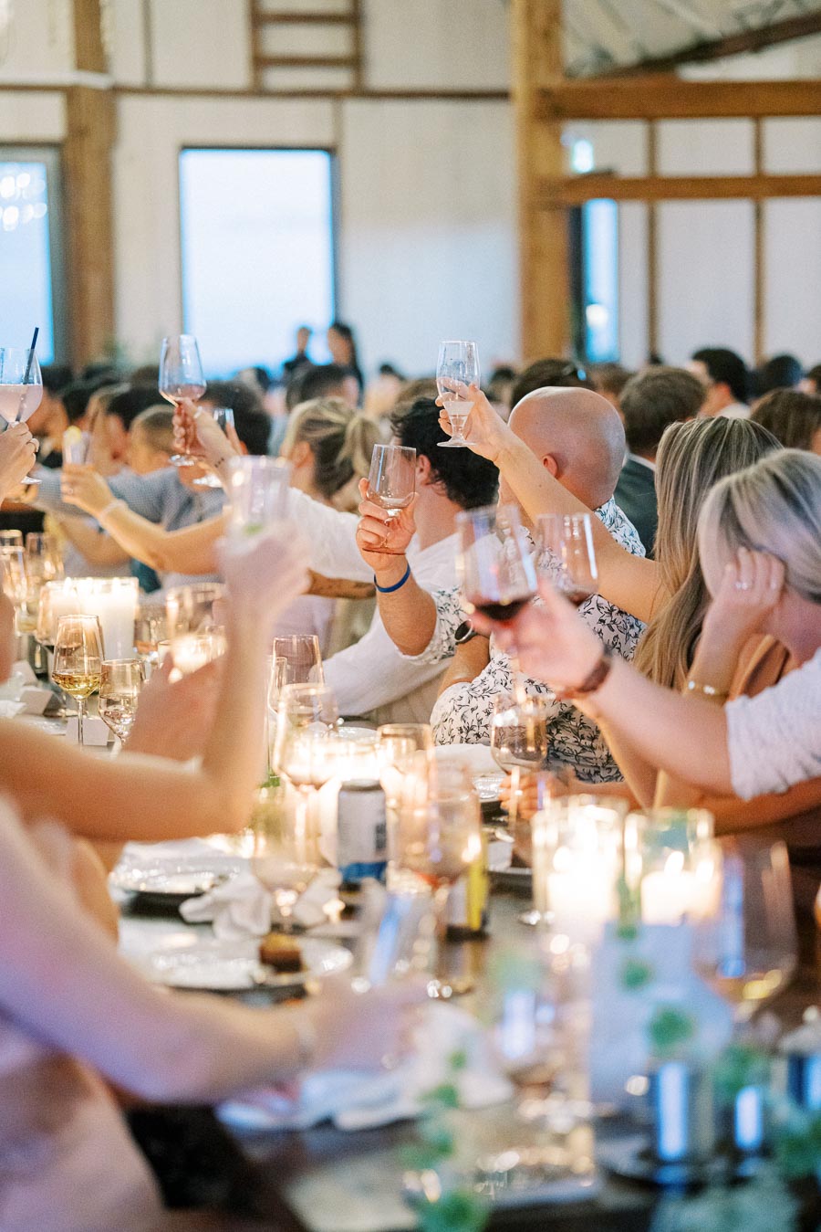 A lively group of people raising glasses in a toast at a beautifully decorated indoor event, with candles and table settings creating an elegant atmosphere.