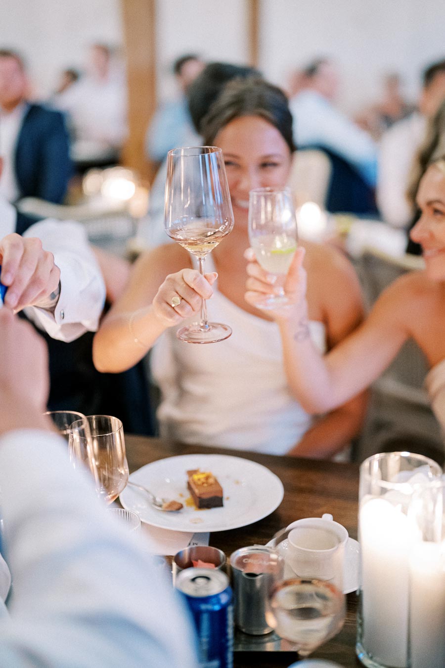 Two women clinking wine glasses in a festive celebration, with a dessert plate and tea candles on the table at a social gathering.