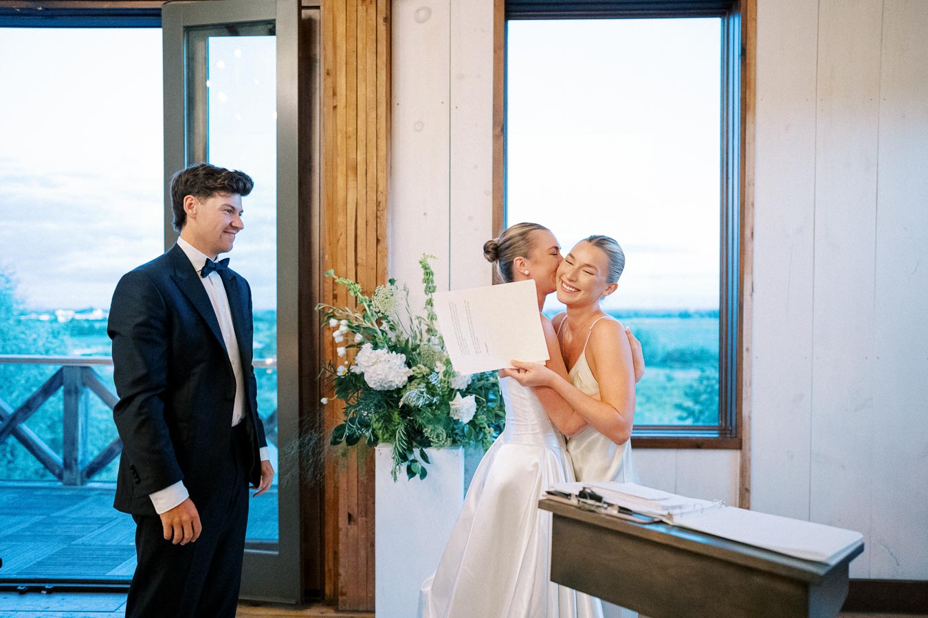 A bride joyfully holds a document while being kissed by another woman in a light-filled room, with a man in a tuxedo smiling nearby. The room features large windows showcasing a scenic outdoor view, enhancing the celebratory atmosphere.