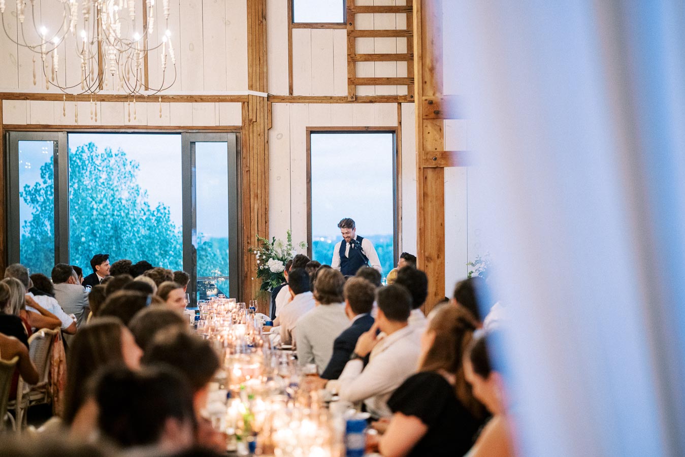 A man gives a speech at a wedding reception in a beautifully decorated rustic venue with chandeliers and large windows, as guests seated at long tables with candlelit centerpieces attentively listen.