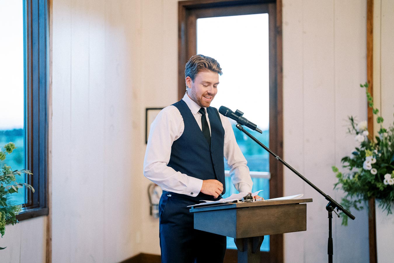 Man in a vest giving a speech at a podium with a microphone in a well-lit room.